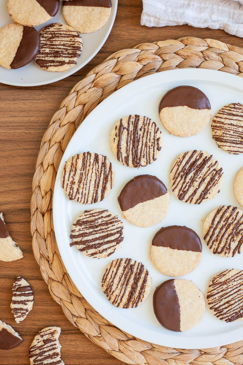 A large white plate is full of thin round cookies placed scattered but not covering each other. Some cookies are partly covered in chocolate, some cookies are drizzled with chocolate so brown stripes can be seen.