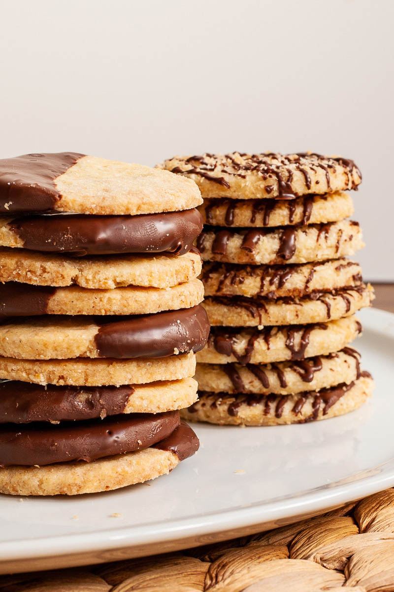 2 stack of thin round cookies on a white plate. One stack has half of the cookies covered with melted chocolate. The other stack has brown stripes of chocolate.