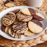A white plate is full of thin round cookies placed placed on top of each other. Some cookies are partly covered in chocolate, some cookies are drizzled with chocolate so brown stripes can be seen.