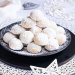A stack of ball-shaped cookies on a black plate. It is dusted generously with powdered sugar so they are white as snow balls. White and silver christmas ornaments are placed next to them.