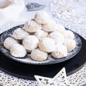 A stack of ball-shaped cookies on a black plate. It is dusted generously with powdered sugar so they are white as snow balls. White and silver christmas ornaments are placed next to them.
