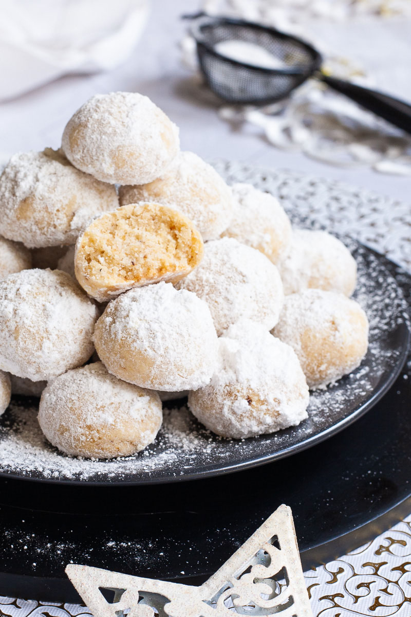 A stack of ball-shaped cookies on a black plate. It is dusted generously with powdered sugar so they are white as snow balls. White and silver christmas ornaments are placed next to them.
