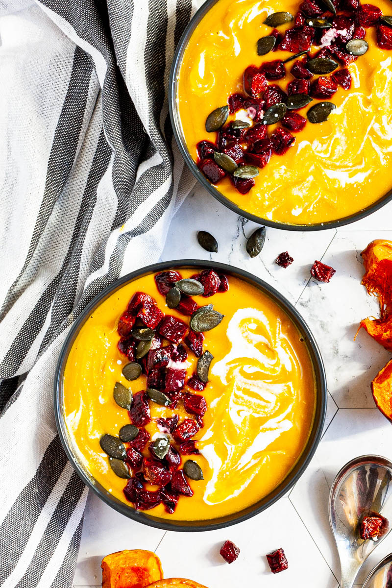 Yellow cream soup in a black bowl topped with diced roasted beetroot, green pumpkin seeds and drizzled with a white sauce.