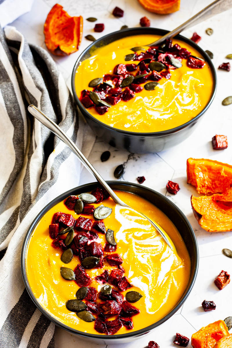 Yellow cream soup in a black bowl topped with diced roasted beetroot, green pumpkin seeds and drizzled with a white sauce.