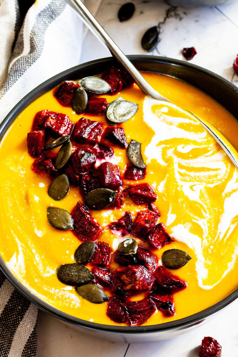 Yellow cream soup in a black bowl topped with diced roasted beetroot, green pumpkin seeds and drizzled with a white sauce.