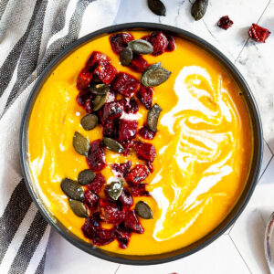 Yellow cream soup in a black bowl topped with diced roasted beetroot, green pumpkin seeds and drizzled with a white sauce.