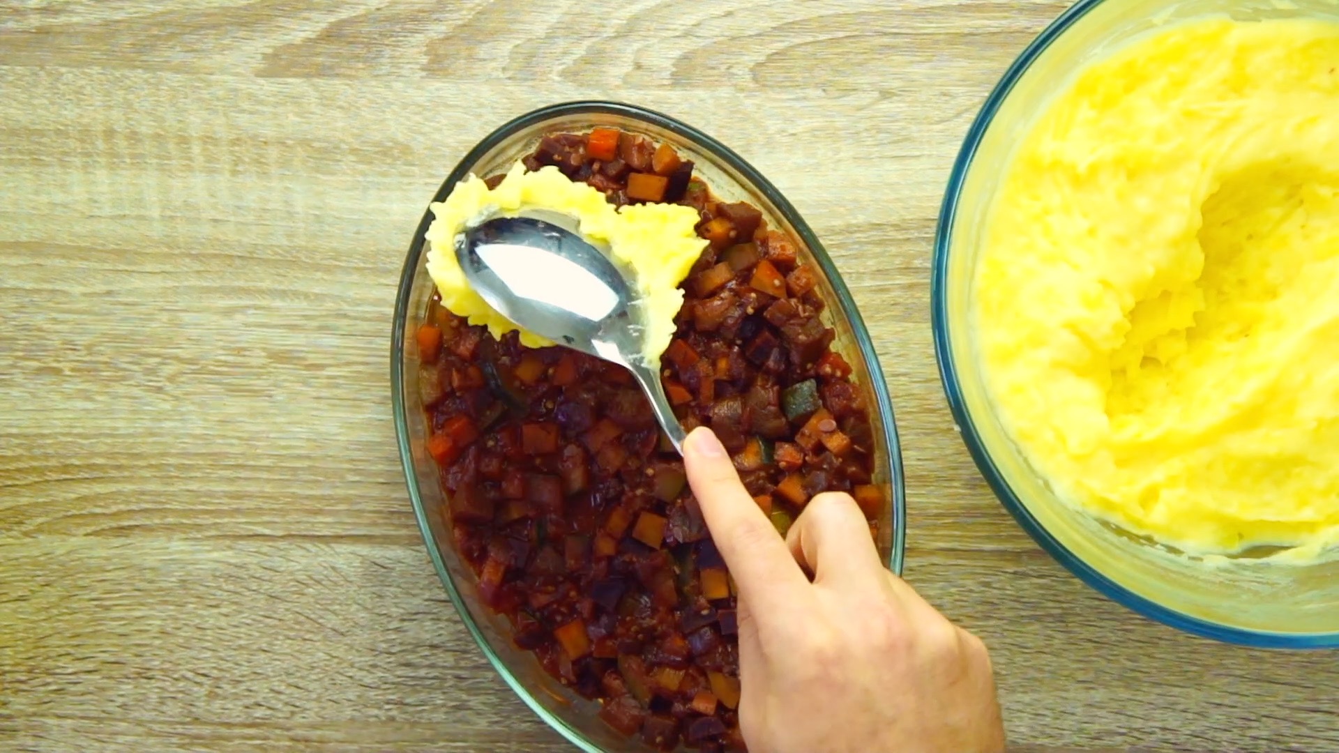 A hand is holding a spoon and covering a veggie stew with mashed potatoes. 