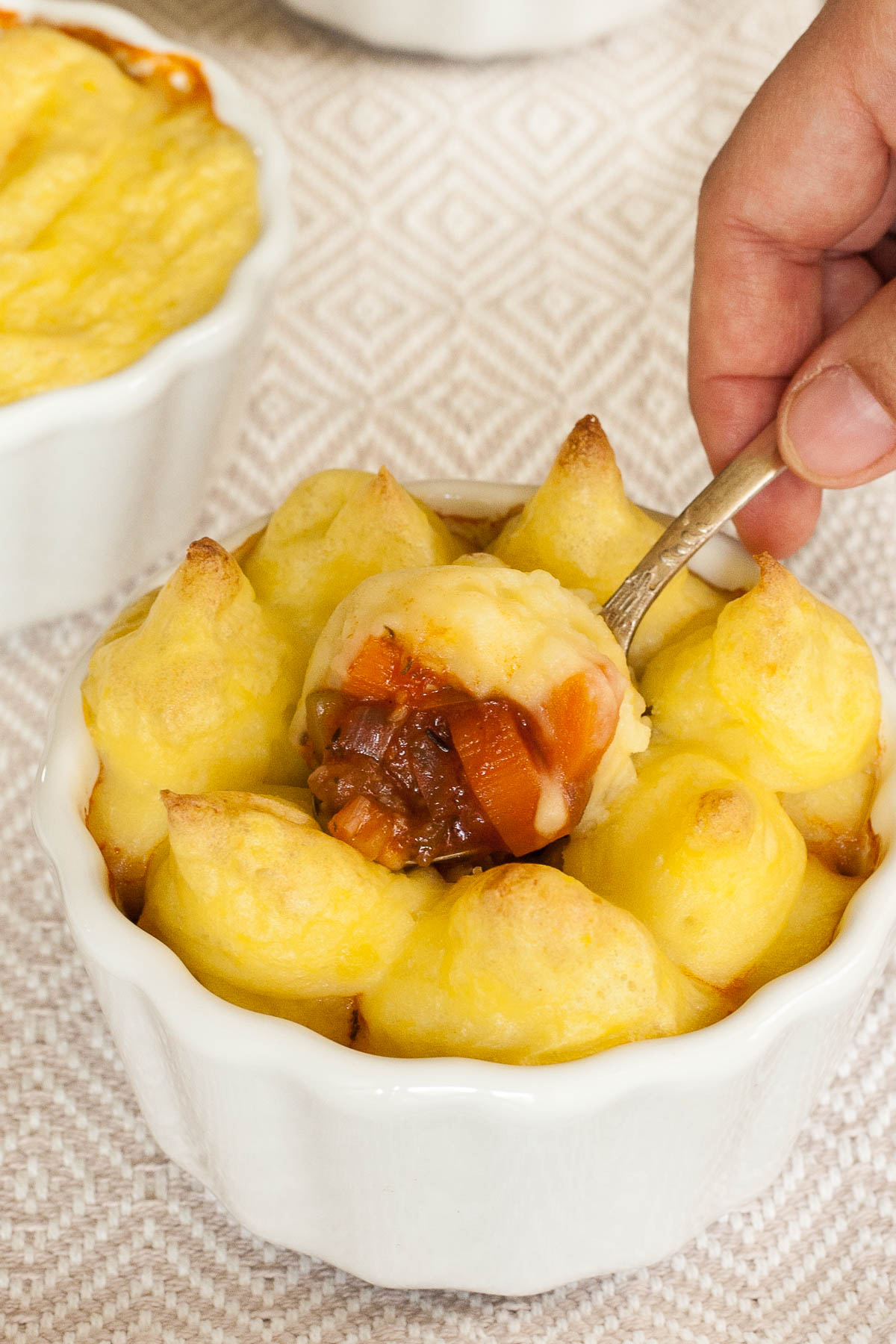 A hand is holding a spoon and takes some from the mashed potato topping and from the veggie stew below out of a white pot of shepherd's pie.