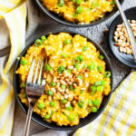 Two black bowls from above on a grey surface full of yellow risotto topped with pine nuts and chopped spring onion. Forks are placed on them.