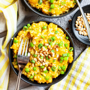 Two black bowls from above on a grey surface full of yellow risotto topped with pine nuts and chopped spring onion. Forks are placed on them.