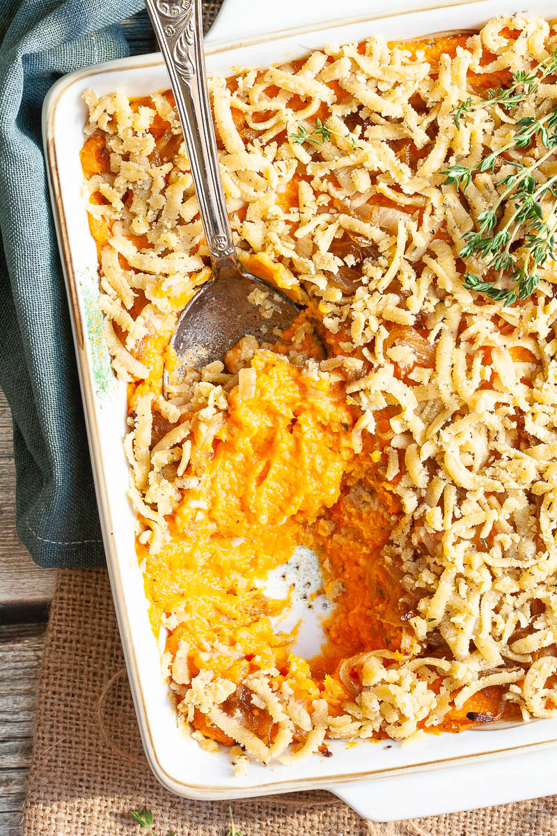 A white casserole dish from above showing orange mash topped with grated crumbles and thyme twigs. A serving spoon is dipped within.