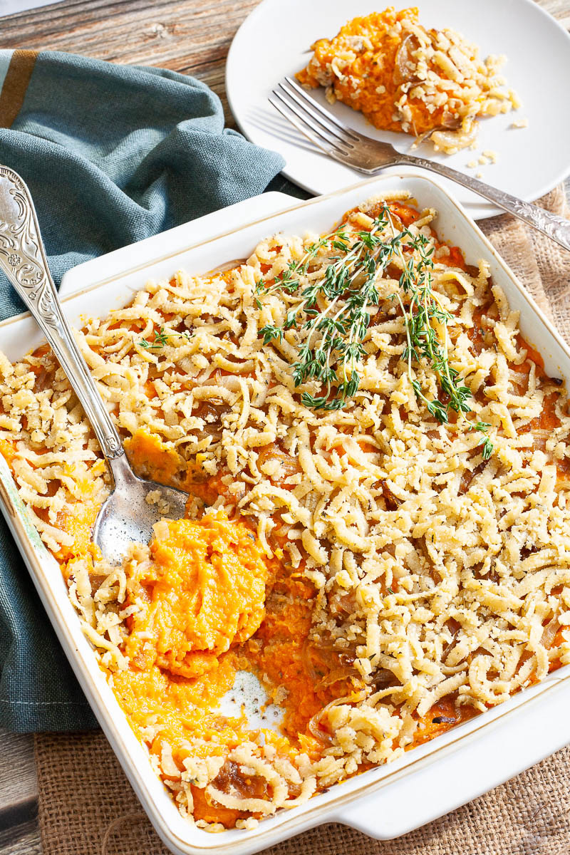 A white casserole dish from above showing orange mash topped with grated crumbles and thyme twigs. A serving spoon is dipped within. The first serving is on the background on a white plate.
