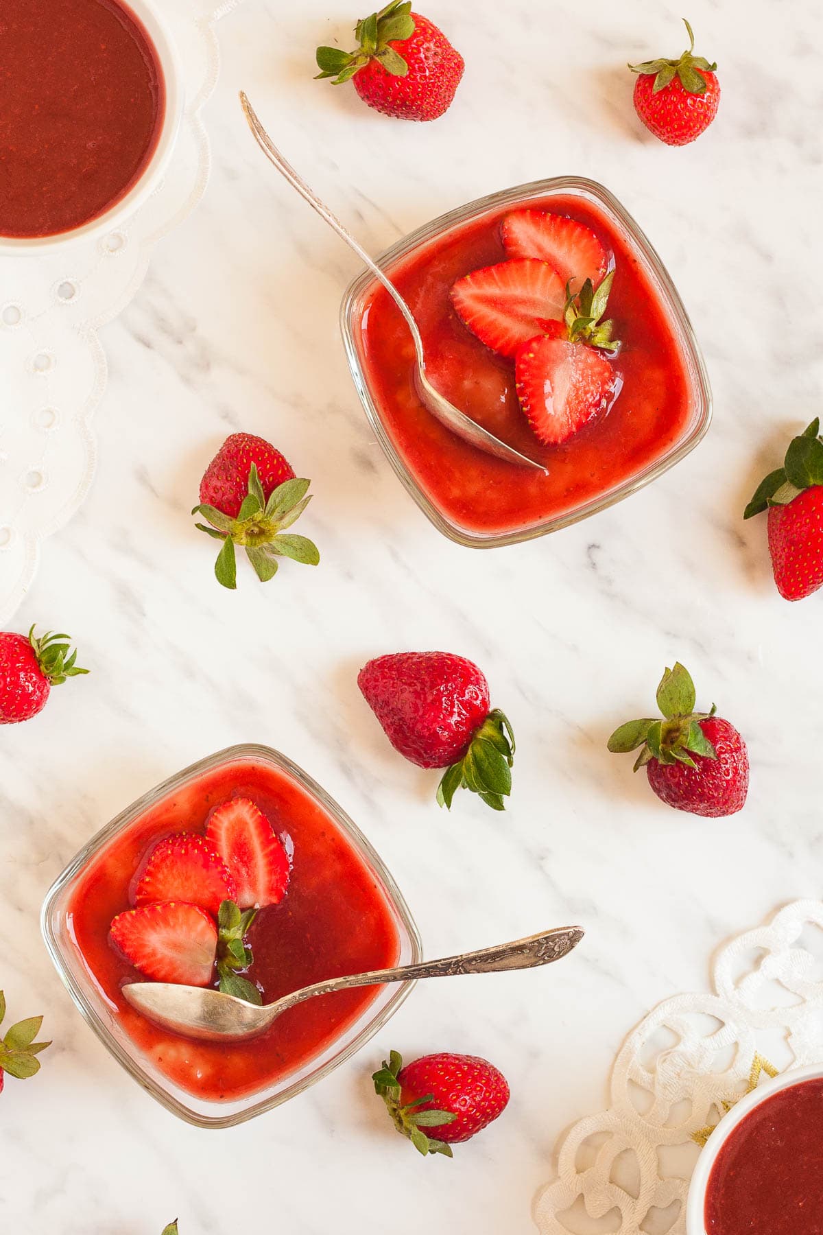 2 small glass bowls with white pudding with pearls topped with strawberry sauce and fresh strawberry slices. Both bowls have spoons. Fresh strawberries are scattered around them.