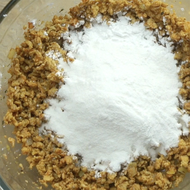 A glass bowl with light brown crumbles mixed with chopped onions and spices. White flour is added on top.