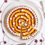 Round tart from above with vibrant yellow filling and pomegranate seeds in a spiral pattern and small whipped cream puffs around and in between. The tart is placed on a white plate and festive decoration is around it.