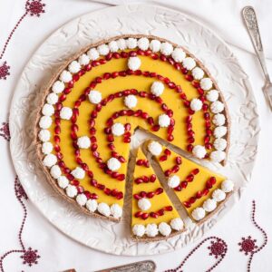 Round tart from above with vibrant yellow filling and pomegranate seeds in a spiral pattern and small whipped cream puffs around and in between. The tart is placed on a white plate and festive decoration is around it.
