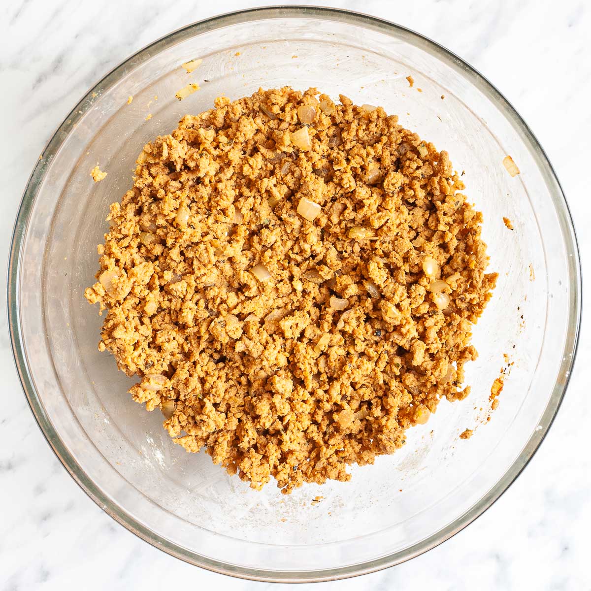 A glass bowl with light brown crumbles mixed with chopped onions and spices.