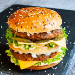 A burger with layers of green ruffled lettuce, yellow sauce, thin cheese slices, dark brown burger patty twice, sliced pickles and white chopped onion.