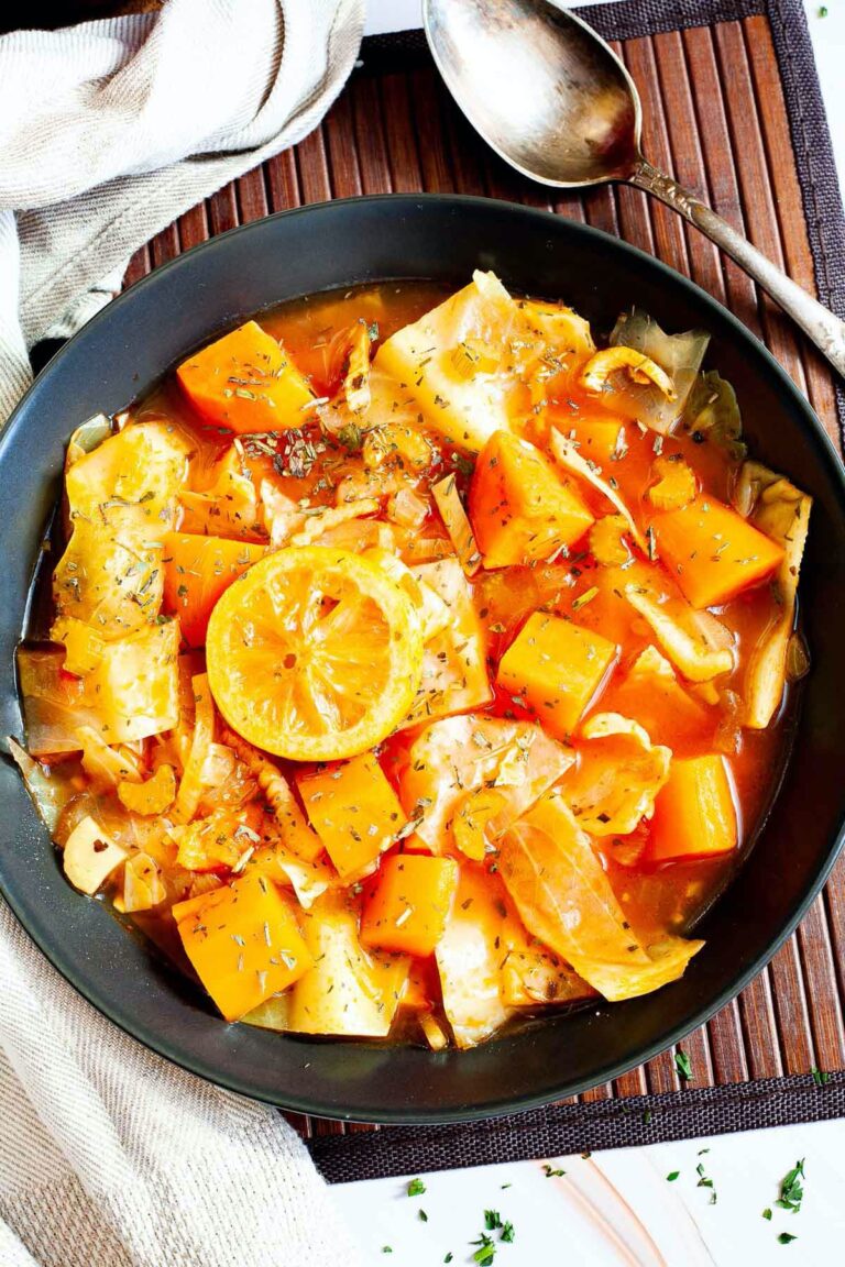 Black plate from above with red tomato-based soup and chopped vegetables like cabbage, orange sweet potato, celery stalk slices, topped with lemon slices and dried green herbs