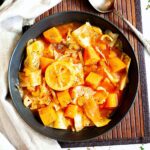 Black plate from above with red tomato-based soup and chopped vegetables like cabbage, orange sweet potato, celery stalk slices, topped with lemon slices and dried green herbs