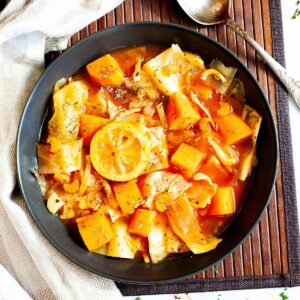 Black plate from above with red tomato-based soup and chopped vegetables like cabbage, orange sweet potato, celery stalk slices, topped with lemon slices and dried green herbs