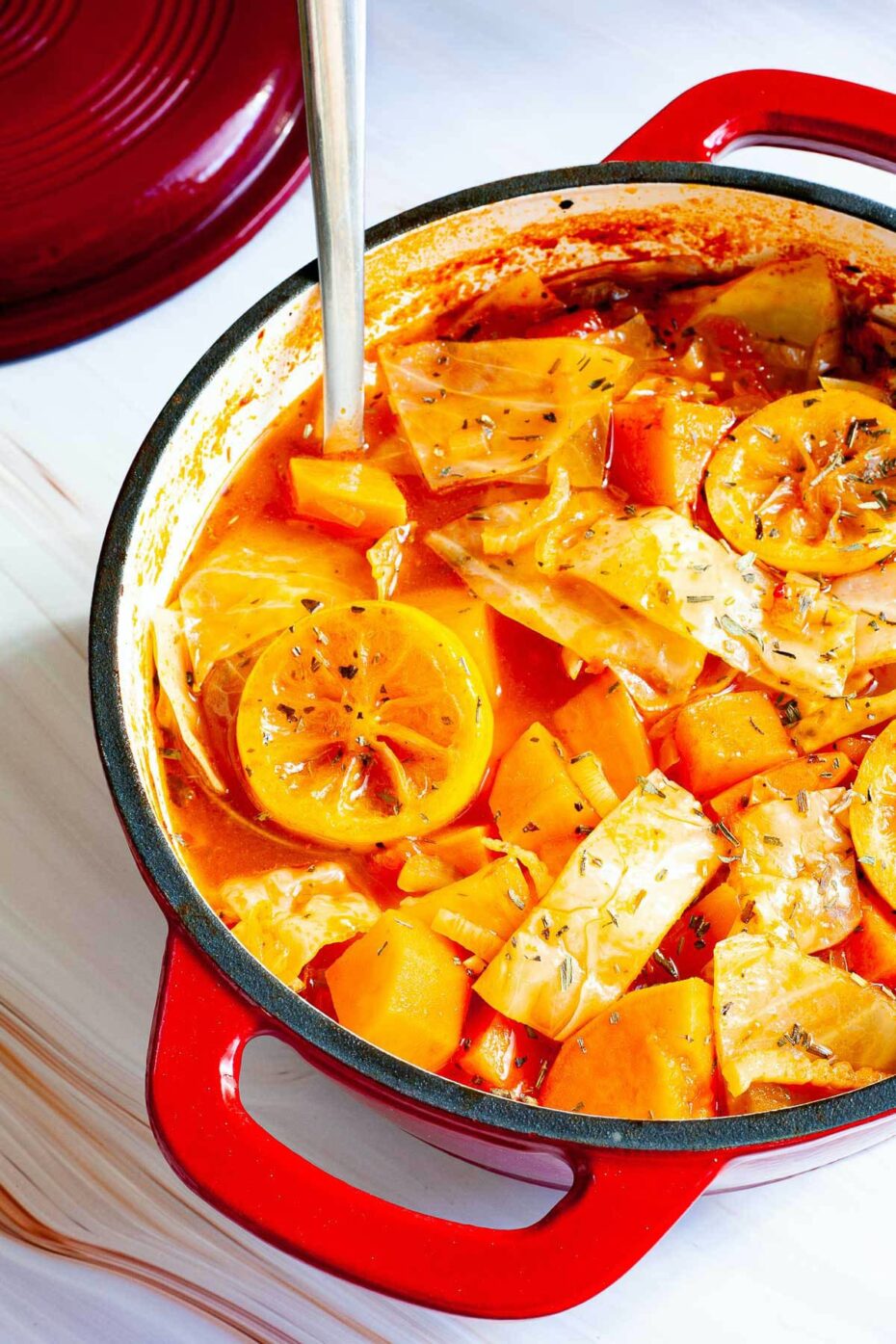 Red dutch oven and ladle with red tomato-based soup and chopped vegetables like cabbage, orange sweet potato, celery stalk slices, topped with lemon slices and dried green herbs