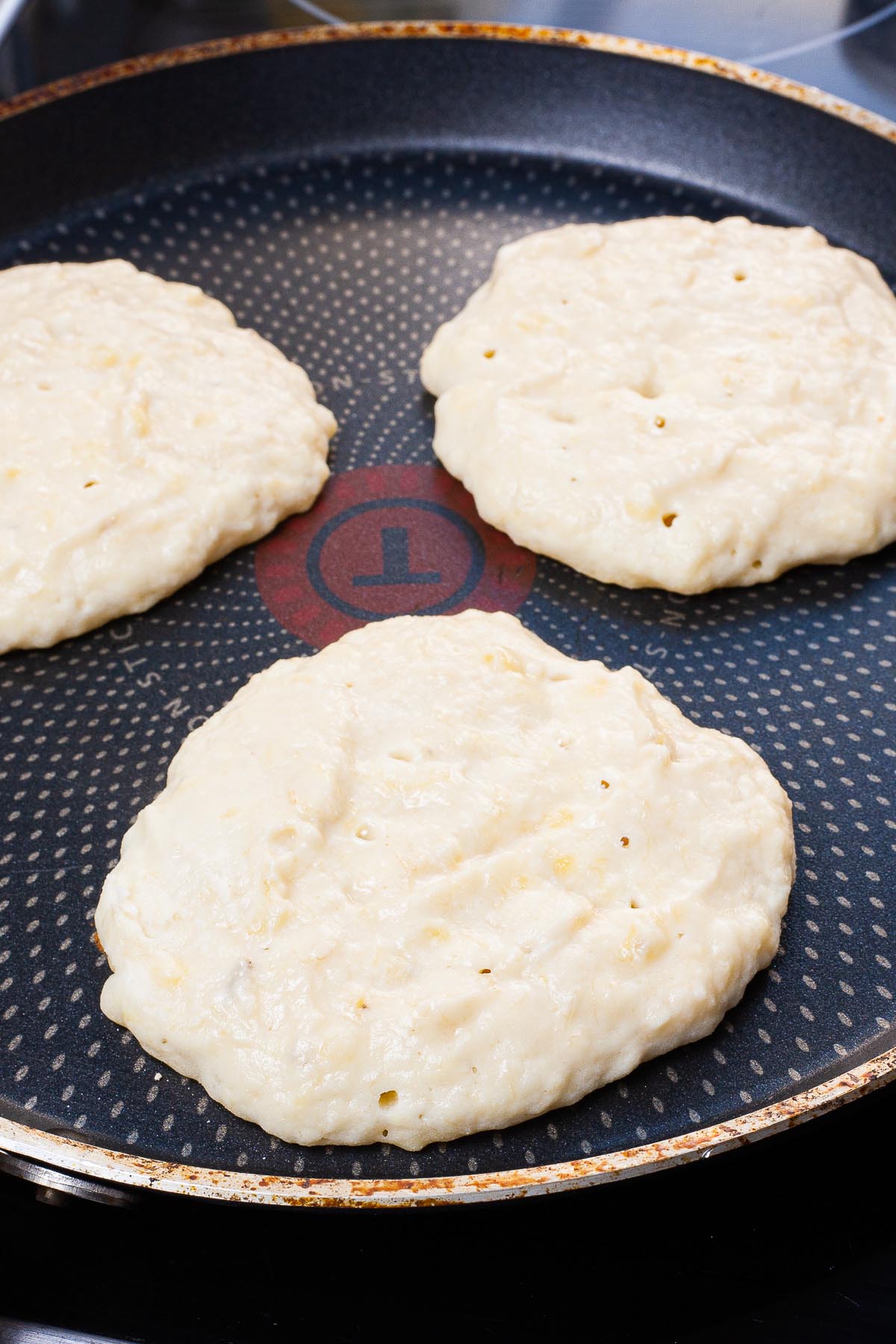 Black frying pan with 3 pancakes that are not yet flipped so the top side is uncooked still