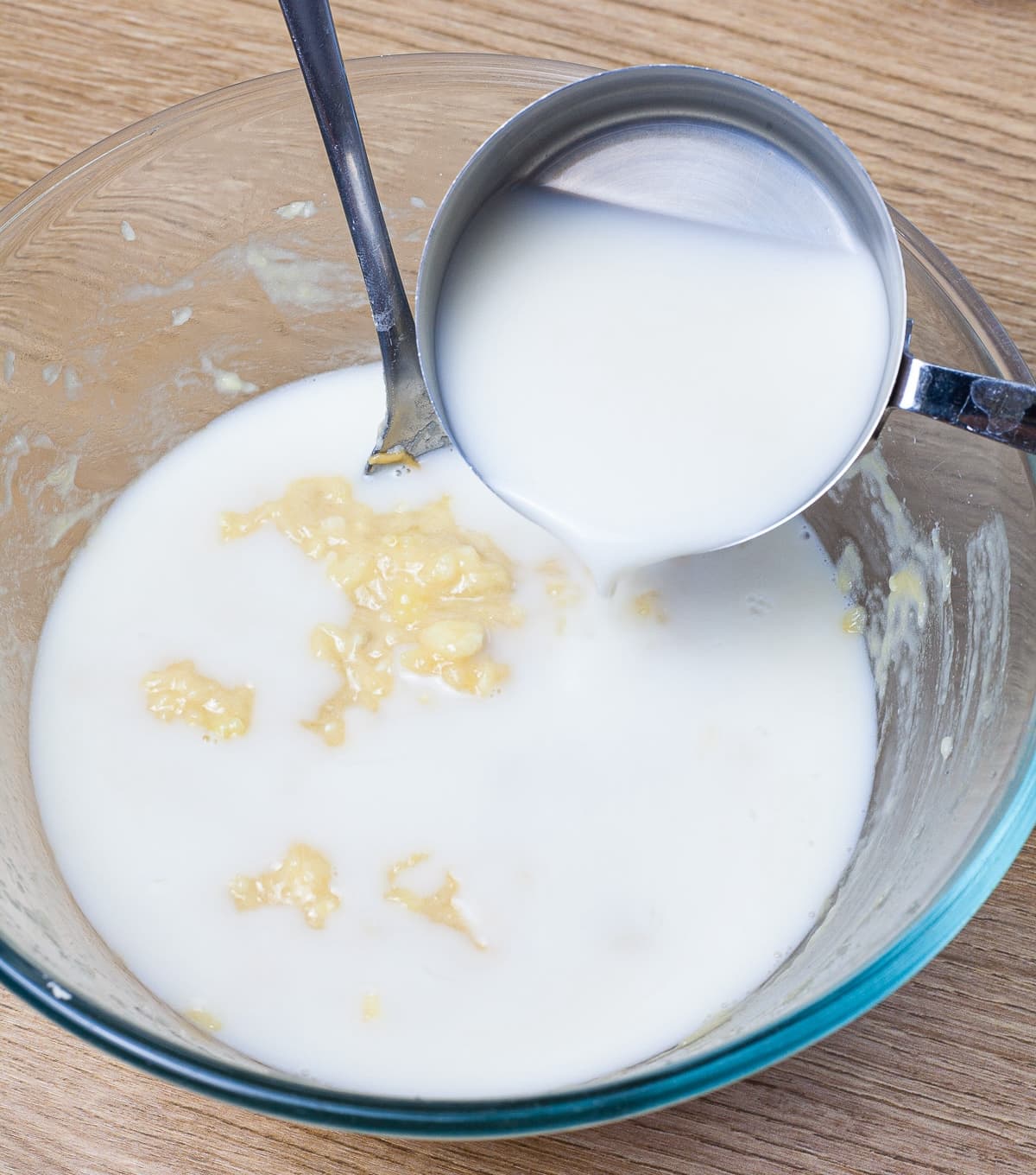 A glass bowl with smashed bananas and a milk is poured from a measuring cup on top of it.