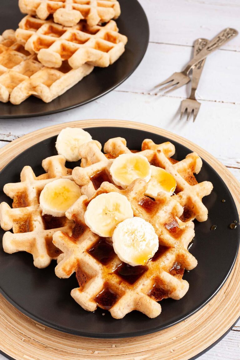 Black plate with a stack of waffles topped with sliced banana and maple syrup. There are 3 waffles on another black plate in the background.