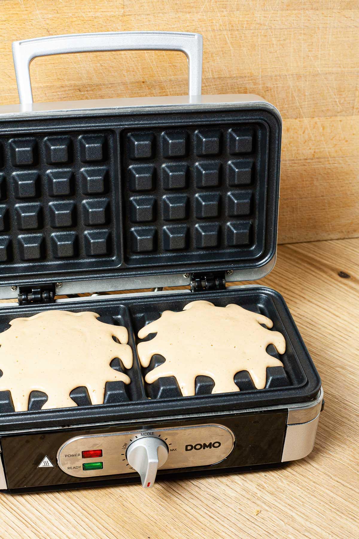 A waffle iron with yellow uncooked batter with a lid open.