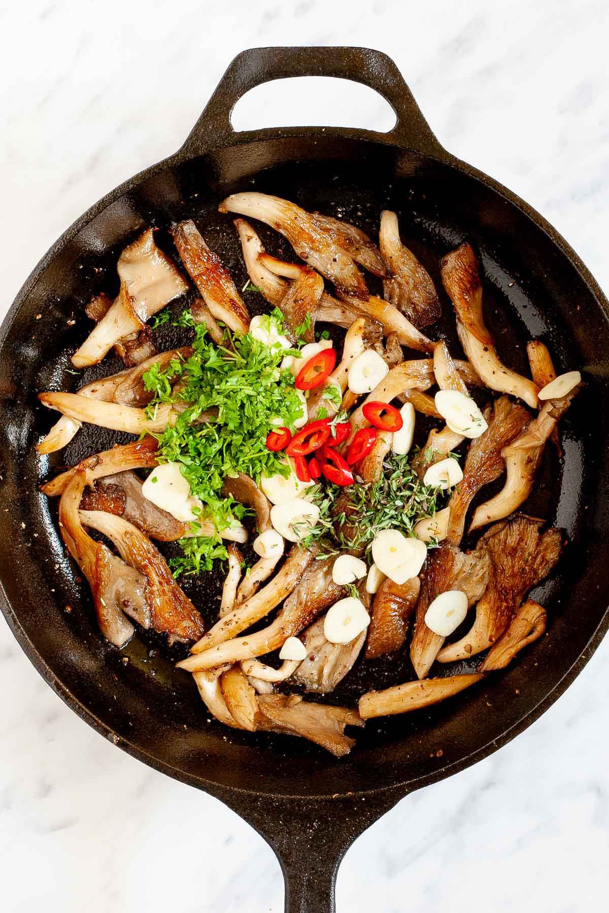 Black cast iron skillet from above with crispy brown oyster mushroom, sliced red pepper, garlic slices and chopped fresh green herbs in groups as they have been just added to the pan.