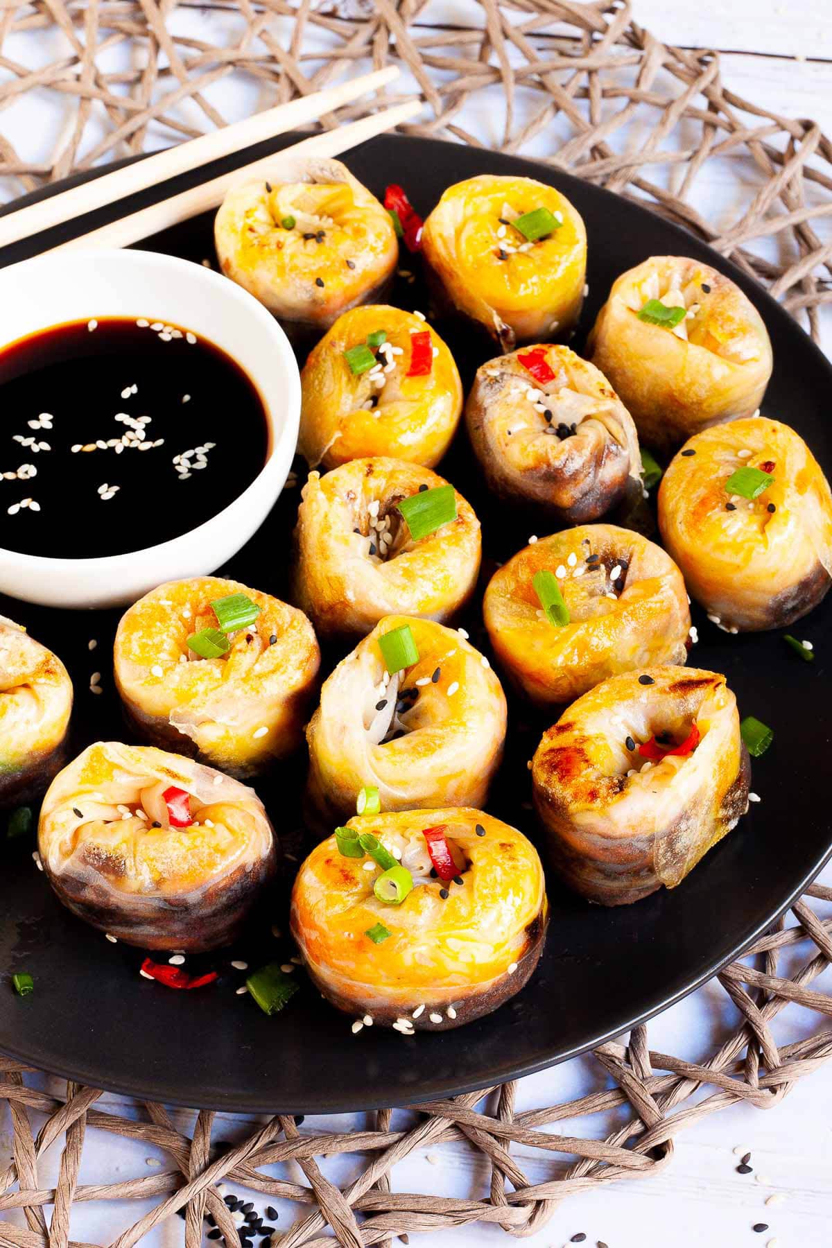 Lots of round-shaped rice paper dumplings arranged in a semi-circular dish facing upwards sprinkled with sesame seeds, chopped spring onion and red hot pepper around a brown dipping sauce.