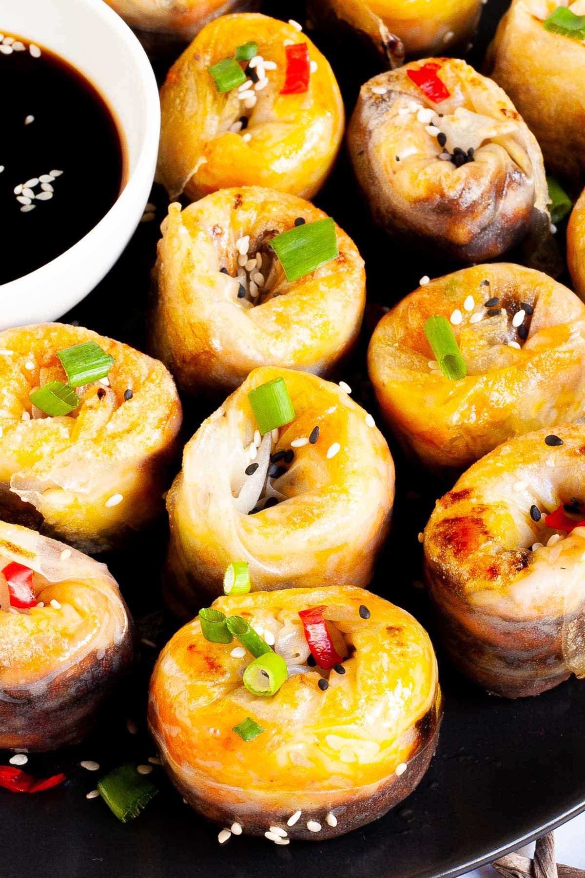 Lots of round-shaped rice paper dumplings arranged in a semi-circular dish facing upwards sprinkled with sesame seeds, chopped spring onion and red hot pepper around a brown dipping sauce.