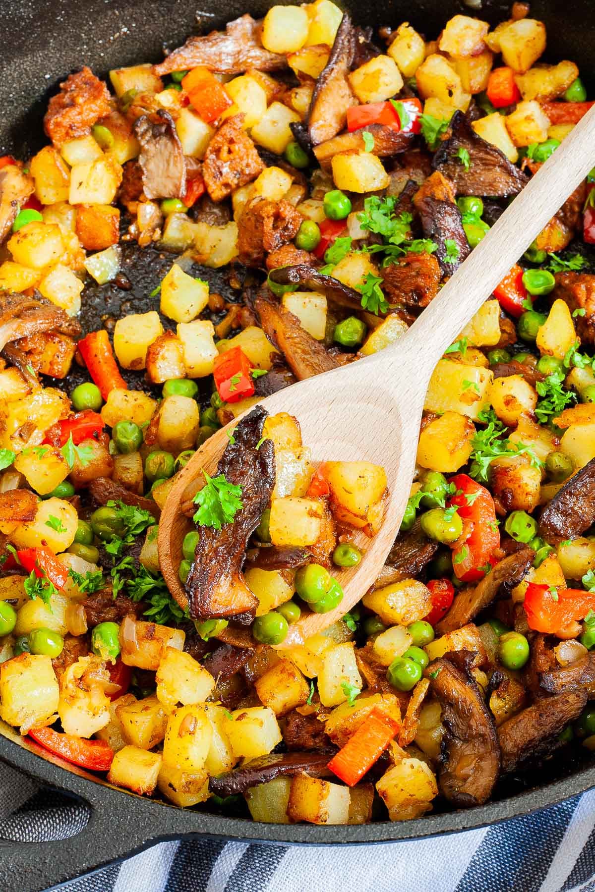Black cast iron skillet from above with a wooden spatula and colorful chopped veggies like red bell pepper, yellow potatoes, brown mushroom, green peas, fresh parsley