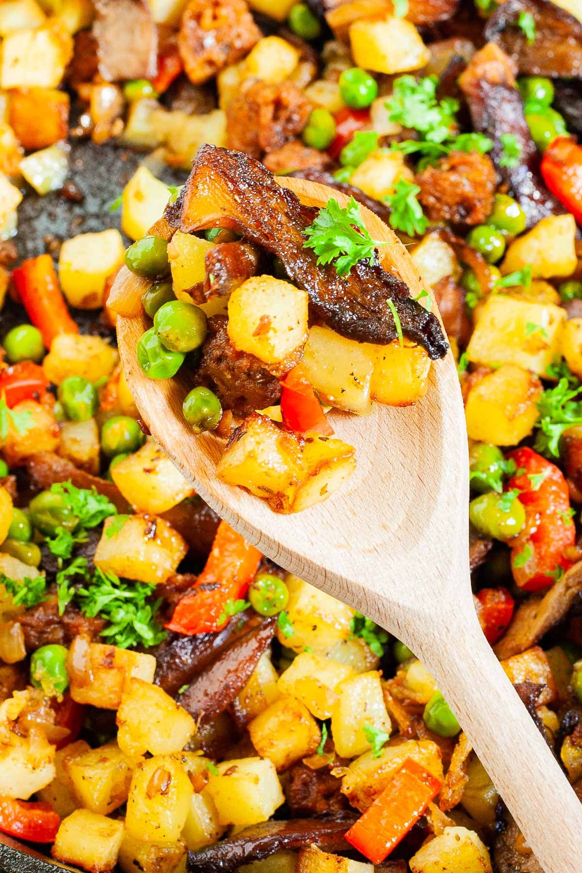 A wooden spoon with colorful chopped veggies like red bell pepper, yellow potatoes, green peas, brown mushroom, fresh parsley.