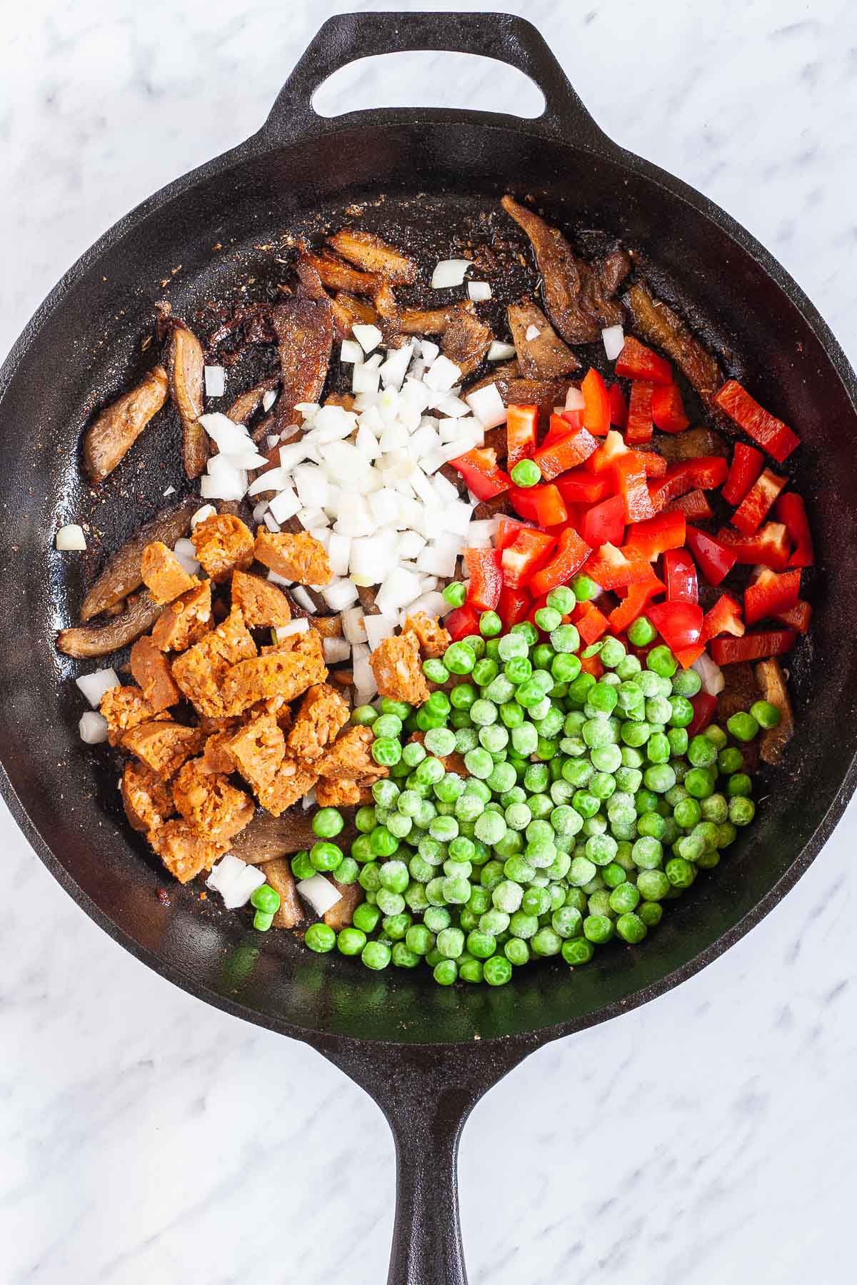 Black cast iron skillet with green peas, chopped red pepper, chopped onion, crispy brown mushrooms and chopped vegan sausage. 