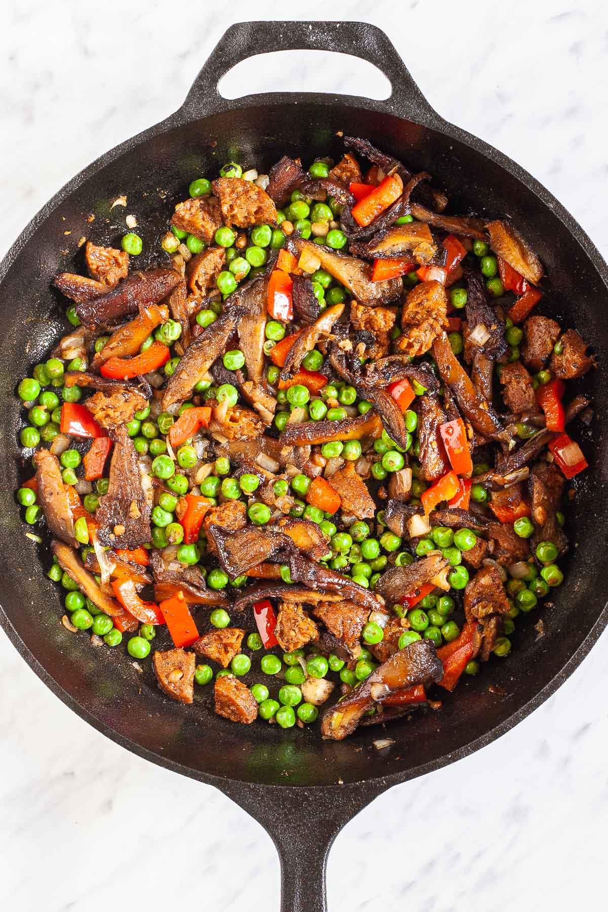Black cast iron skillet with a mix of fried veggies like green peas, chopped red pepper, chopped onion, crispy brown mushrooms and chopped vegan sausage.
