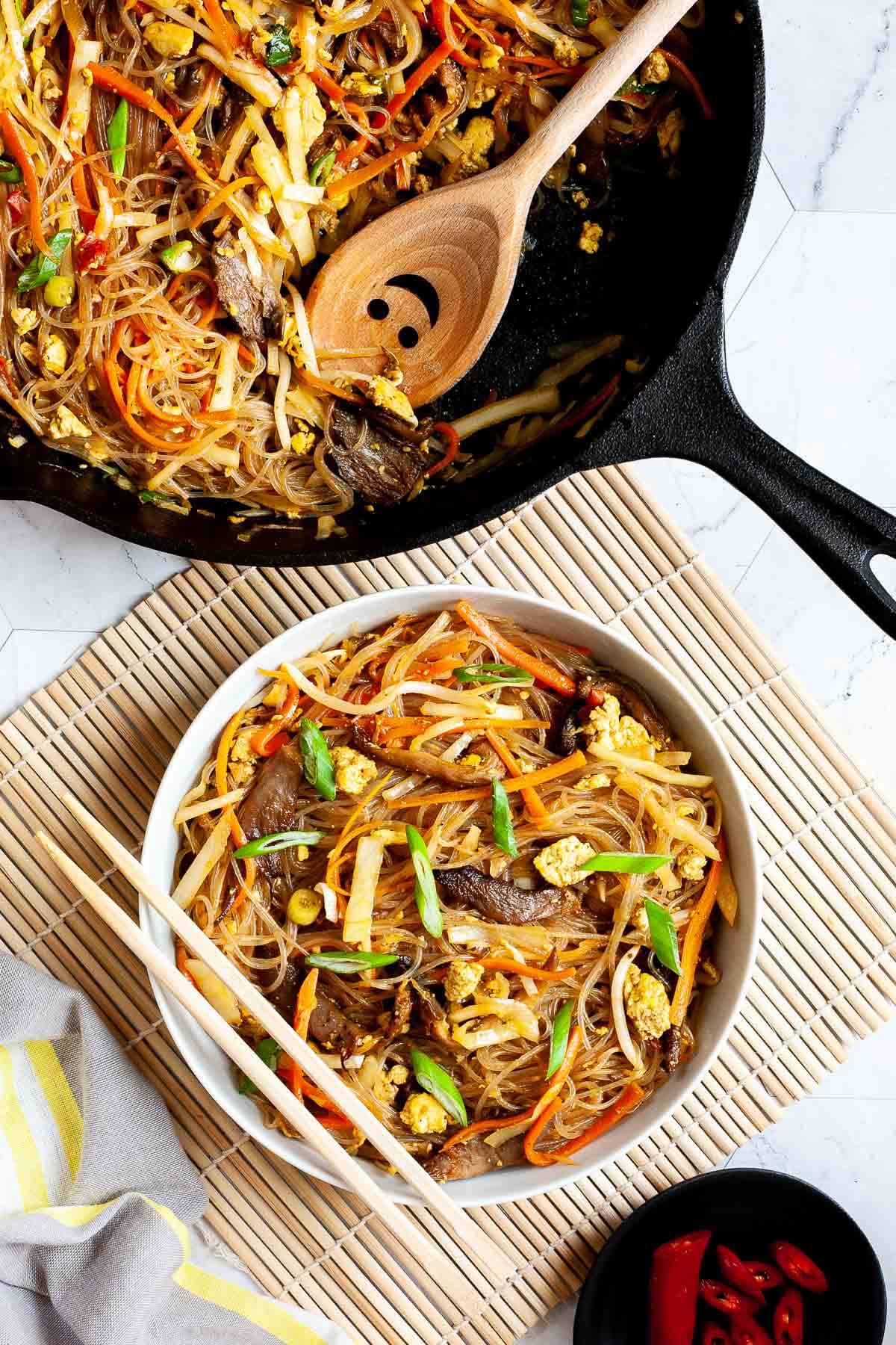 Black skillet from above full of glass noodles, shredded carrots, cabbage, sliced green onion, bean sprouts, mushroom shreds with a smiling wooden spatula. A white serving plate with chopsticks on it have the same contents