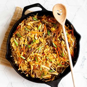 Black skillet from above full of glass noodles, shredded carrots, cabbage, sliced green onion, bean sprouts, mushroom shreds with a smiling wooden spatula