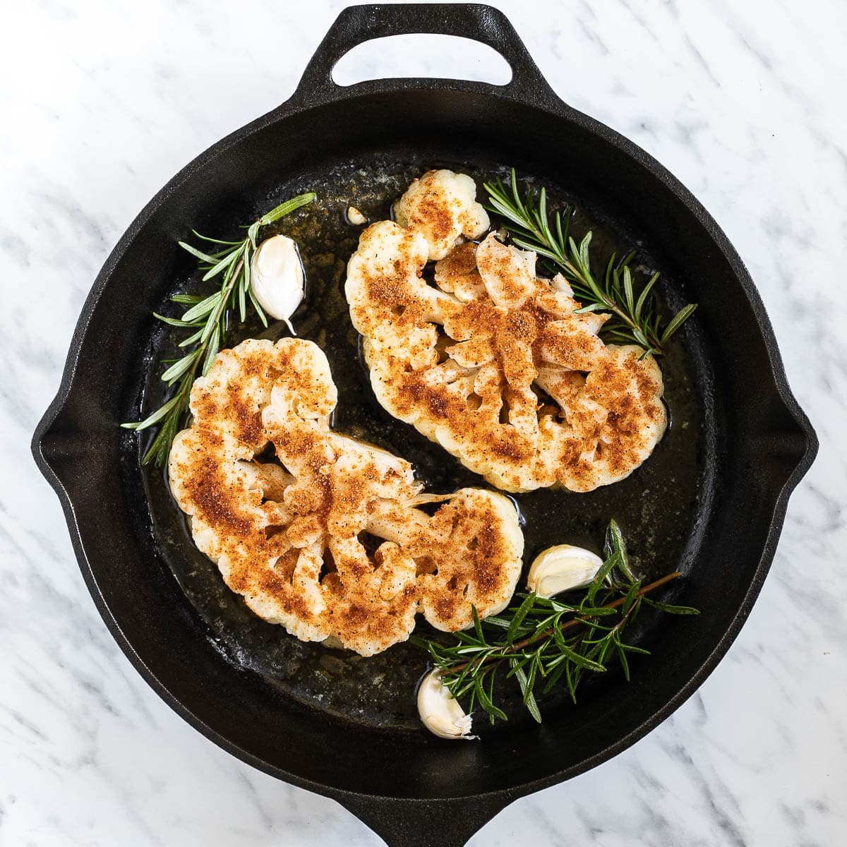 2 heavily spiced cauliflower slices in a cast-iron skillet surrounded by garlic cloves and rosemary twigs.