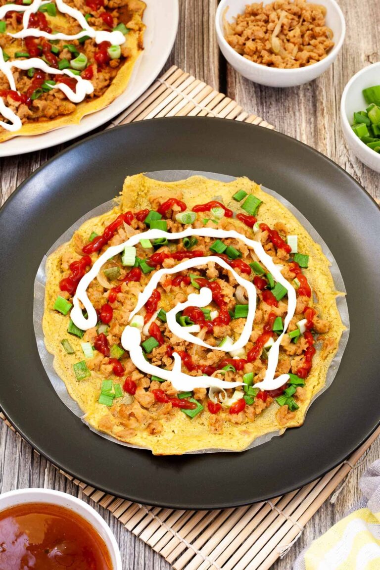 Black plate with opaque rice paper topped with a yellow batter and sprinkled with brown crumbles and green spring onion rings. White and red sauces are drizzled on top.