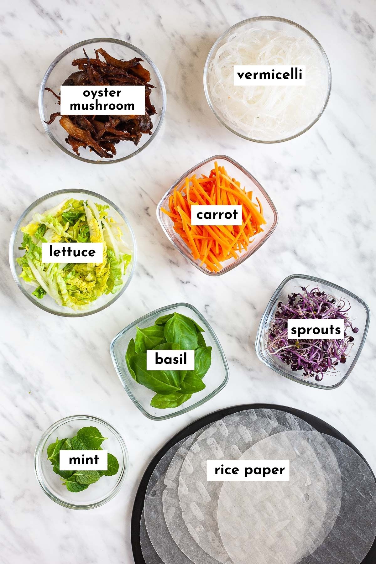 All ingredients of Vietnamese summer rolls are placed in small glass bowls like brown mushroom slices, glass noodles, lettuce, carrot, basil leaves, mint leaves, purple sprouts. Dry rice paper sheets are on a black plate. 