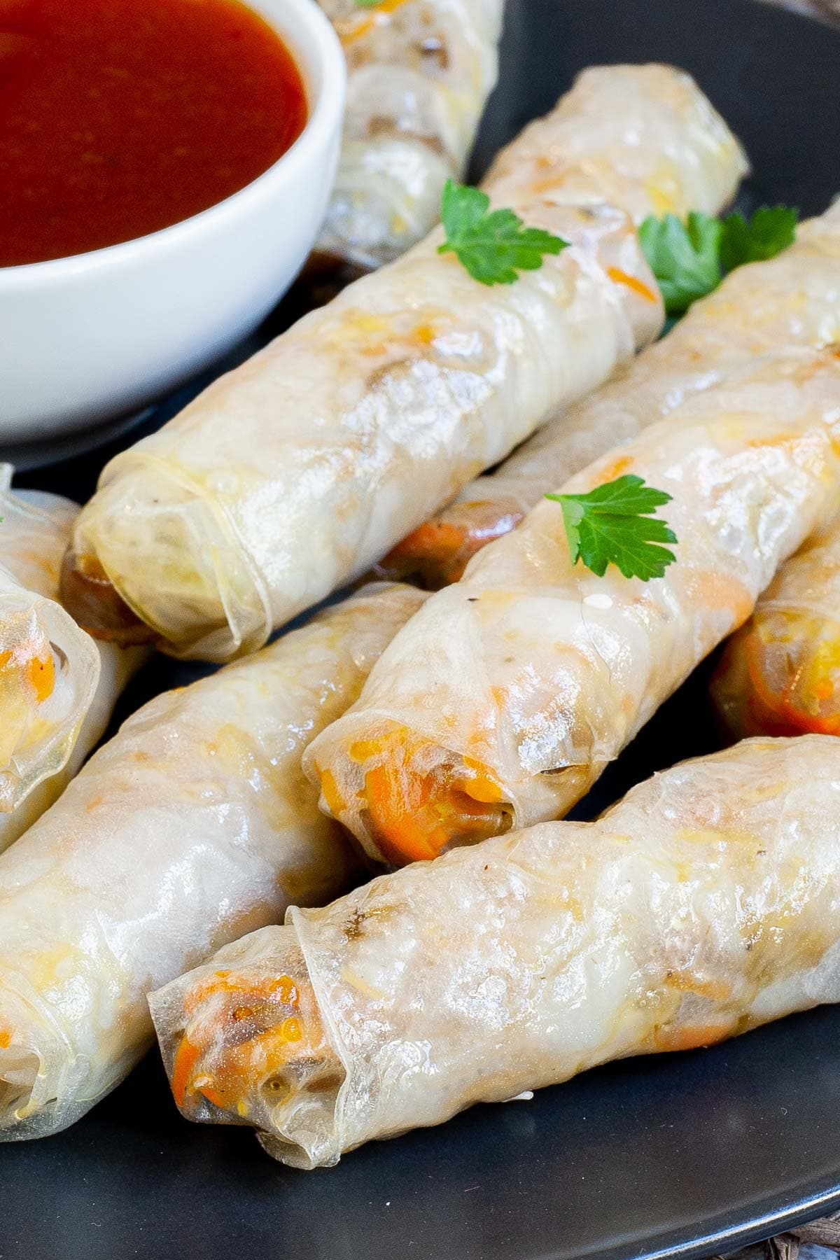 Black plate with crispy looking light brown egg rolls. A small white bowl is with a red dipping sauce. The rolls are sprinkled with parsley leaves.