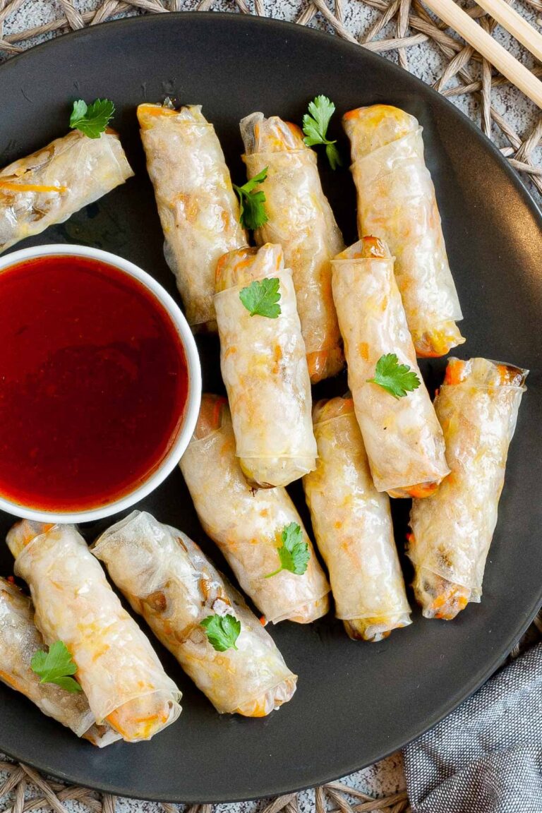 Black plate with crispy looking light brown egg rolls. A small white bowl is with a red dipping sauce. The rolls are sprinkled with parsley leaves.