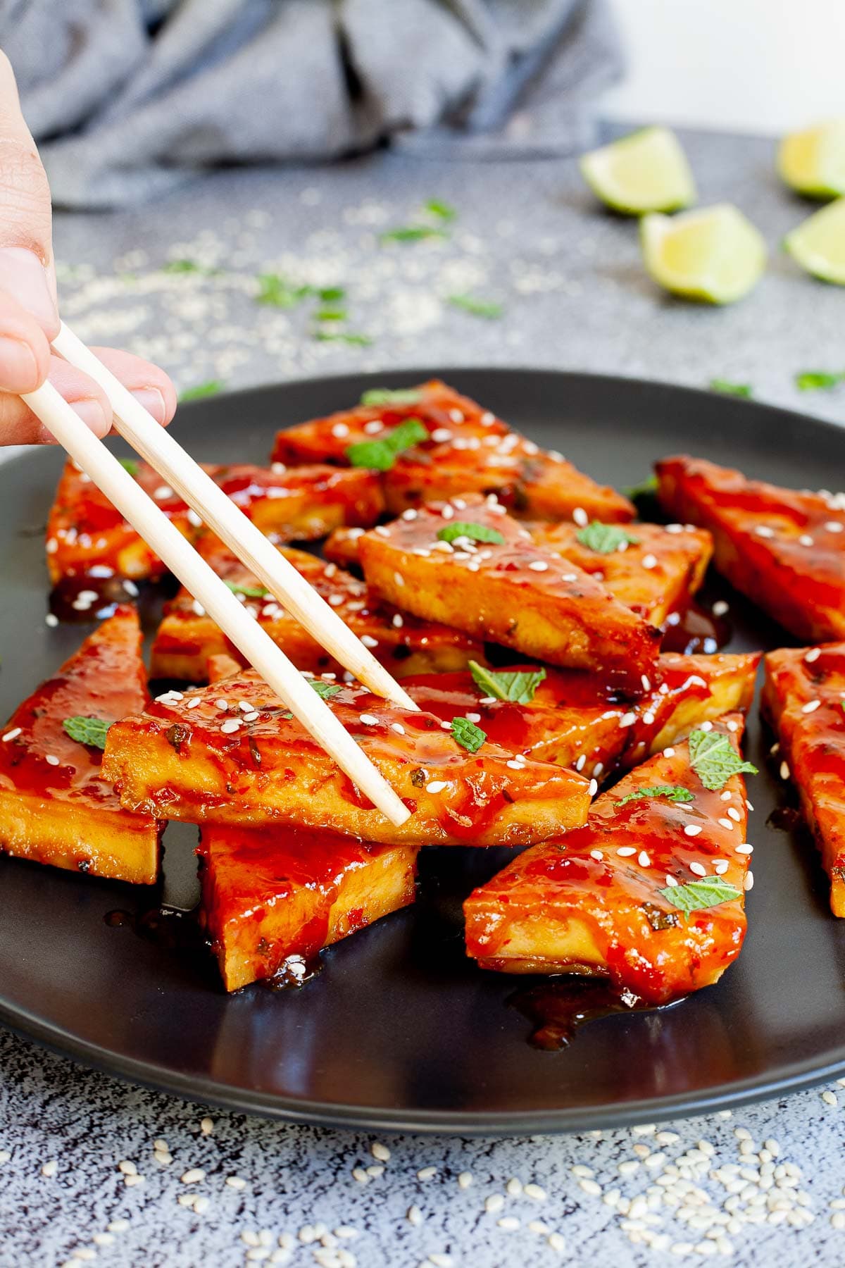 Triangle-shaped tofu slices in a red sticky glaze with freshly chopped green mint, sesame seeds on black plate. A hand with chopsticks is about to take one.