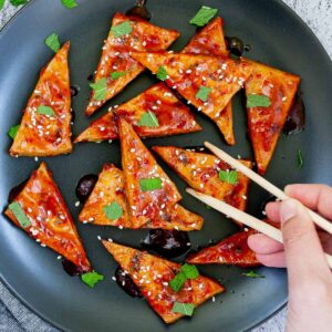 Triangle-shaped tofu slices in a red sticky glaze with freshly chopped green mint, sesame seeds on black plate. A hand with chopsticks is about to take one.