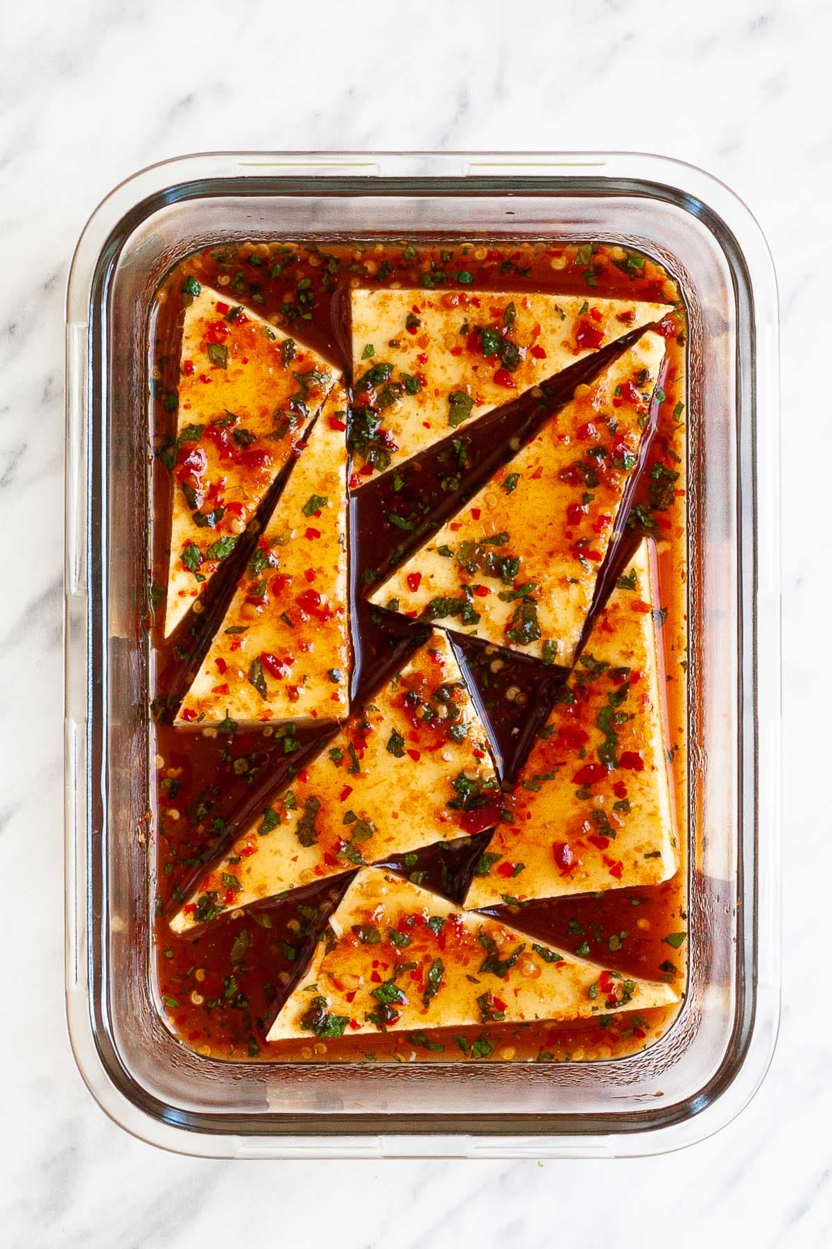 Rectangular glass dish with triangle shaped tofu slices in a red brown liquid marinade with pepper seeds and green chopped leaves.