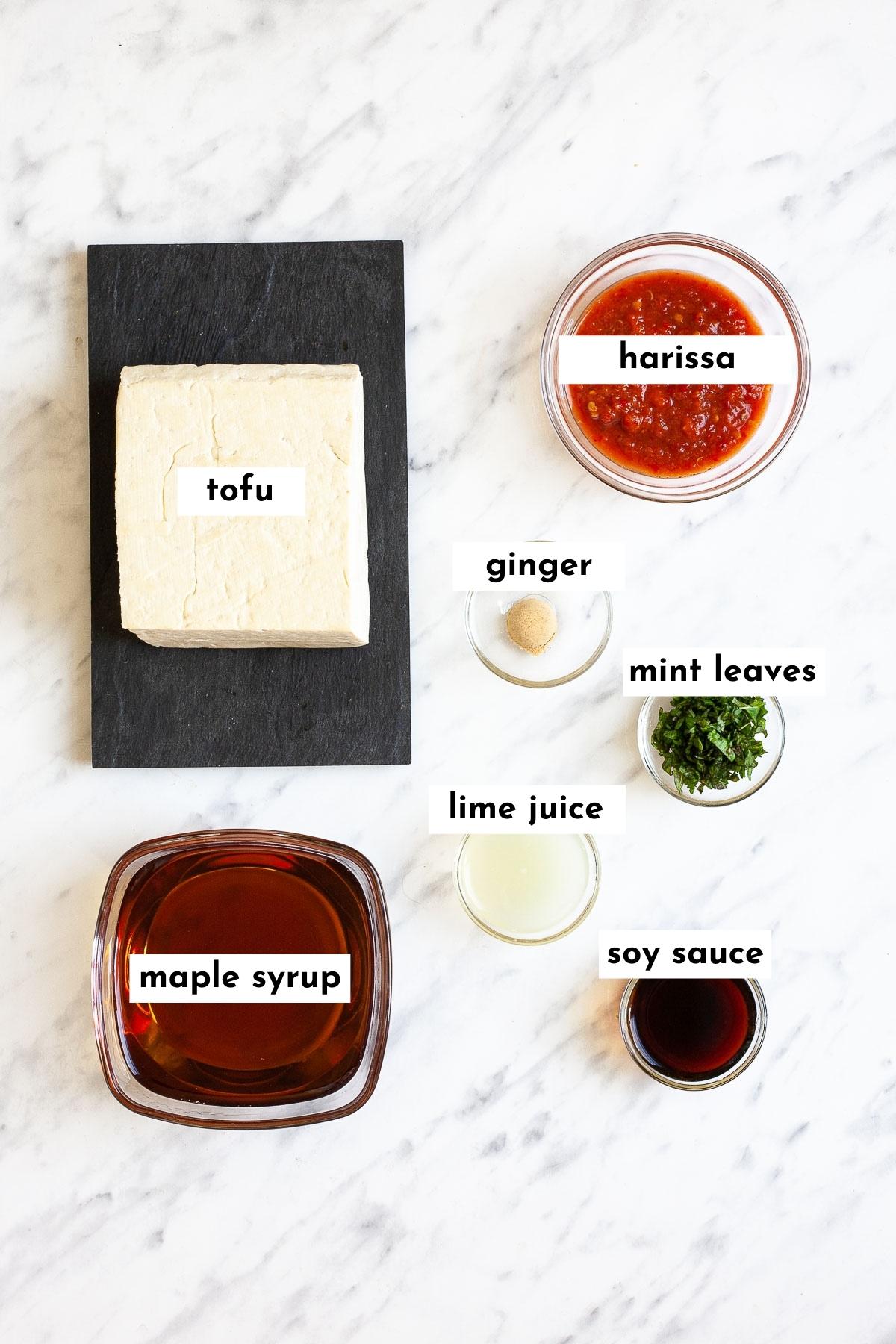 ingredients of harissa tofu in small glass bowls like harissa, maple syrup, lime juice, ginger powder, mint leaves, soy sauce and a block of tofu on a black plate