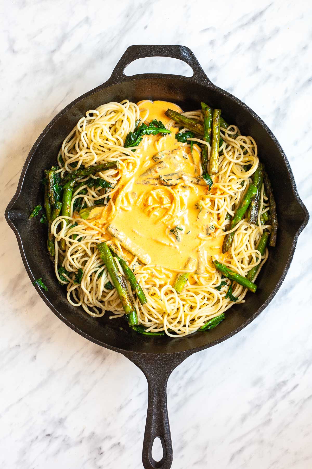 Cast iron skillet with asparagus pieces, spinach leaves, and spaghetti topped with a yellow sauce.