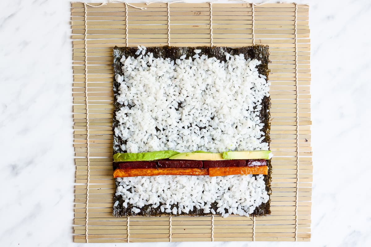 Bamboo mat with seaweed sheets, rice, avocado slices, sweet potato and purple beet sticks.